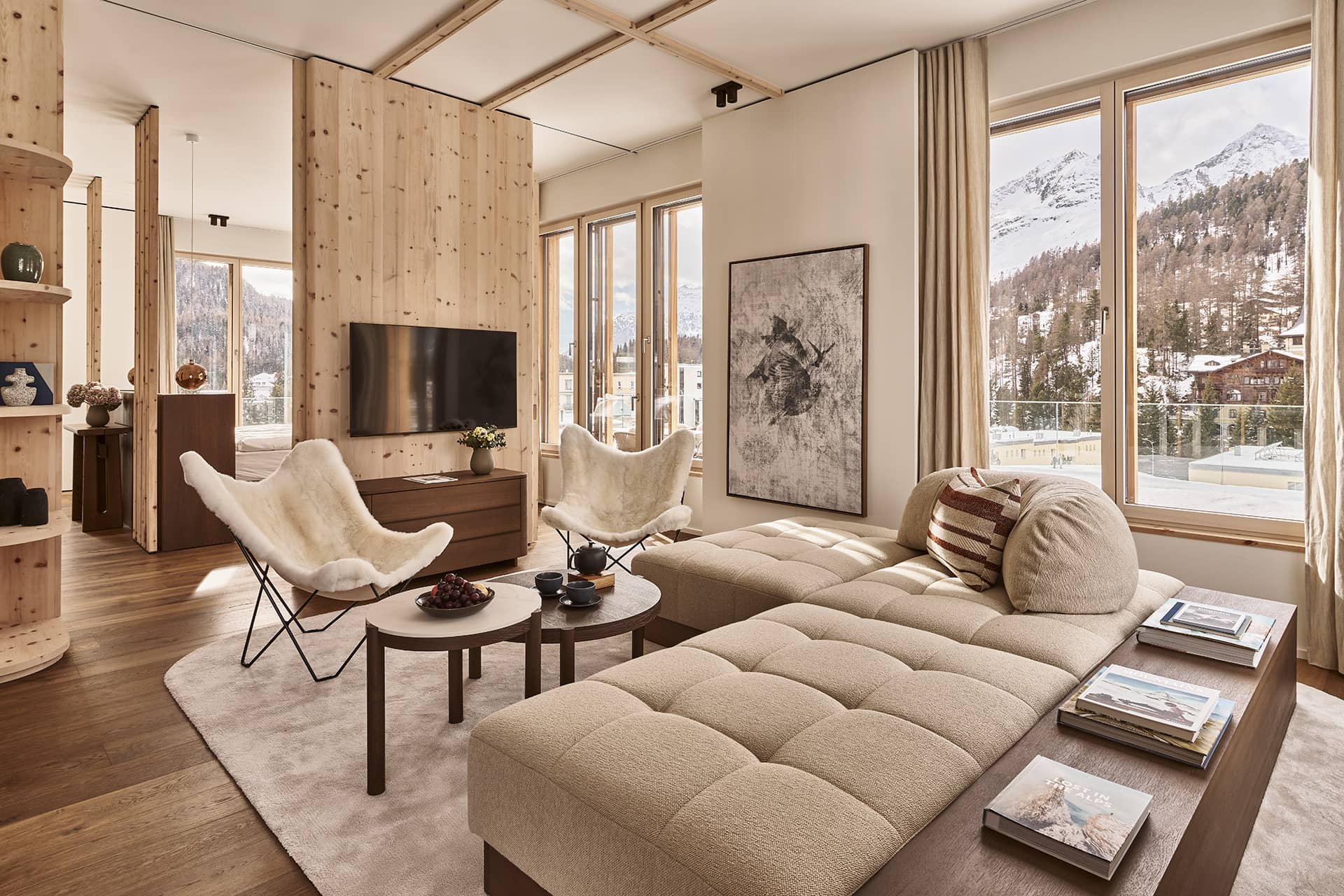 Wohnbereich im alpinen Penthouse mit Panoramafenstern, Bergblick, beigem Modulsofa, zwei Loungesesseln aus Lammfell und Holzvertäfelung.