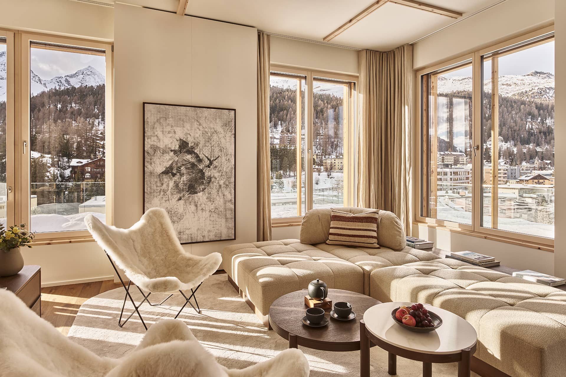 Heller Wohnraum mit großer Fensterfront und Blick auf verschneite Berglandschaft, Sofa in Naturtönen und runden Holztischen.