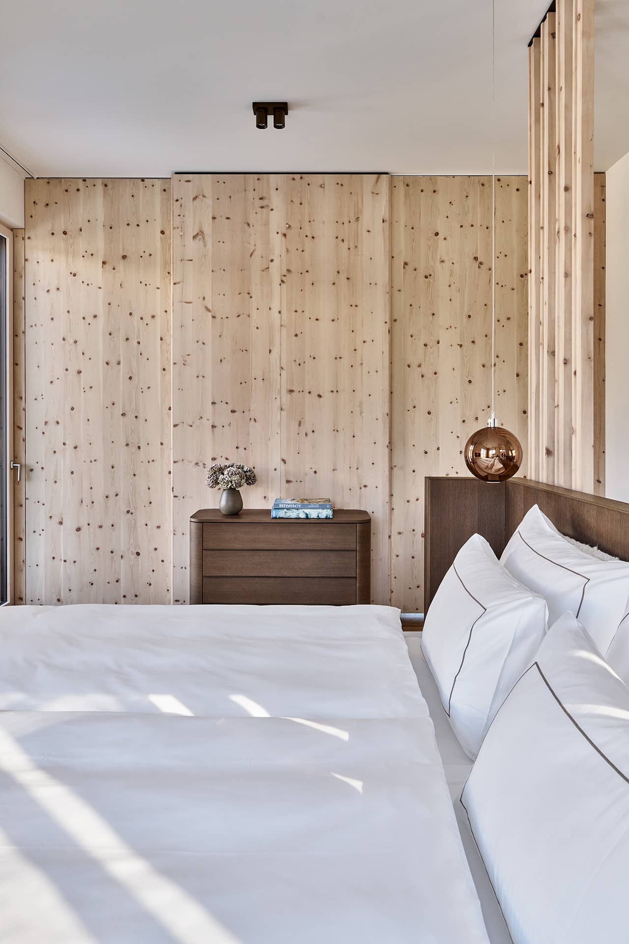Schlafzimmer mit weissem Doppelbett, Holzelementen und Blick in den angrenzenden Wohnbereich.