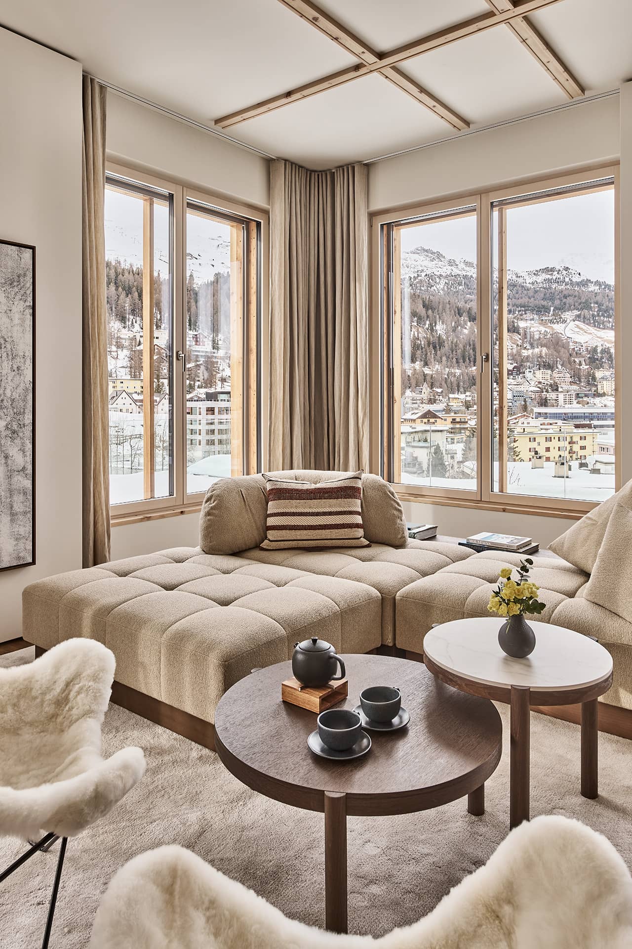 Wohnlounge mit beigem Sofa und runden Couchtischen vor grossen Eckfenstern mit Alpenblick.